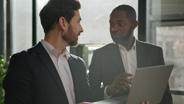 Caucasian man sales manager talking to African American male company client in office. Two diverse multiracial men employees workmates discuss business project software with laptop agree startup idea