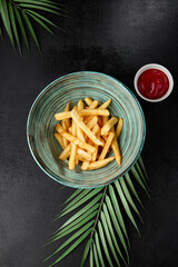 French fries in ceramic bowl on black concrete background. Composition with french fries and palm leaf on dark background. Junk food in modern style. Fried potatoes in asian style.