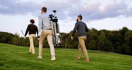 Three men with clubs and golf bag walking on the golf fied. Back view from below. Outside