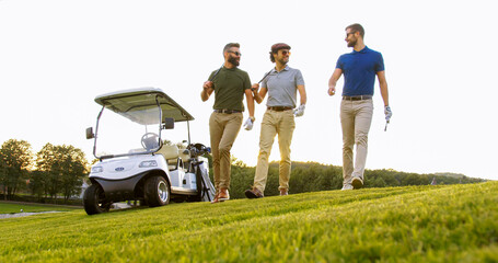 Three handsome men strolling with their clubs and golf bags on the golf field to start a game and talking