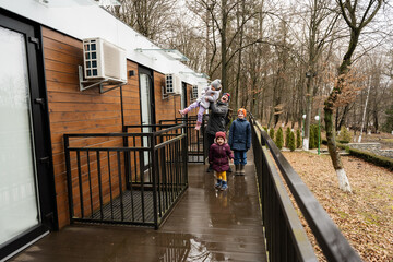 Fototapeta premium Father with three kids on terrace of one-storey modular houses in spring rainy forest.