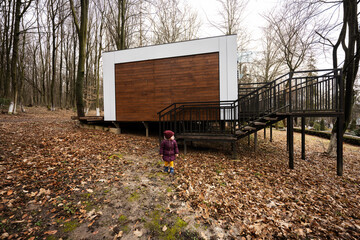 Baby girl against one-storey modular house in spring rainy forest.