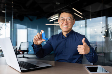Businessman satisfied with asthma medicine inside office, man holding inhaler in hands smiling and looking at camera and showing thumbs up, successful asian man at workplace working with laptop. - obrazy, fototapety, plakaty