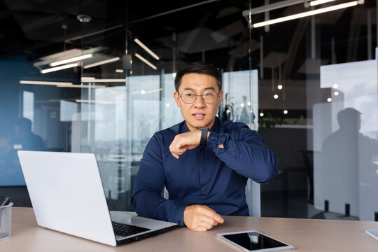 Successful businessman inside office, using app on smart watch to communicate, asian man giving voice commands to artificial intelligence through watch, boss working sitting with laptop.
