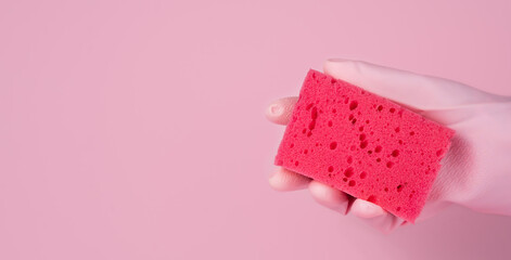 A woman's hand in a rubber glove holds a cleaning sponge on a pink background. Washing and cleaning concept. Close-up. Copy space. Banner. Selective focus.