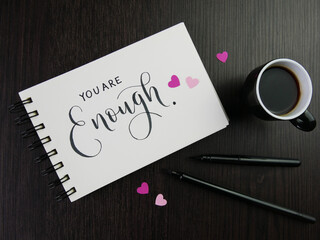 YOU ARE ENOUGH lettering in notepad with cup of coffee and pens on black desk