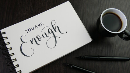 YOU ARE ENOUGH lettering in notepad with cup of coffee and pens on black desk