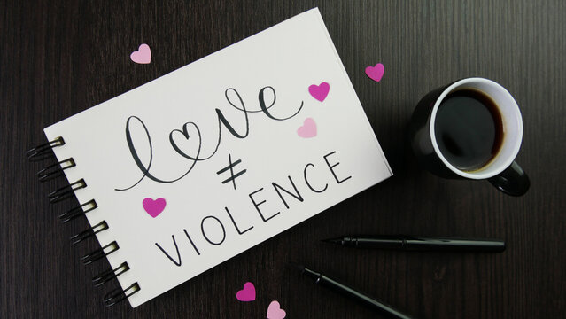 LOVE ≠ VIOLENCE lettering in notepad with pink hearts, cup of espresso and pens on desk