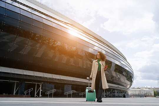 Young african woman walking outdoors carrying a suitcase and going to travel by airplane at modern airport. Vacations, travel and active lifestyle concept