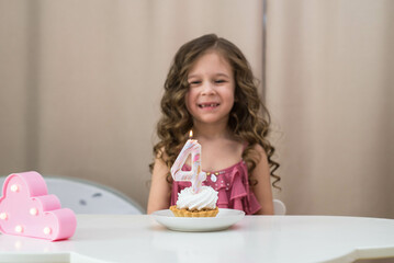 Obraz na płótnie Canvas little girl is smiling and looking at birthday cake on white table in light room.