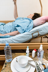 white cup with saucer and medicines on table and sick woman holding handle of homecare hospital bed on background at home