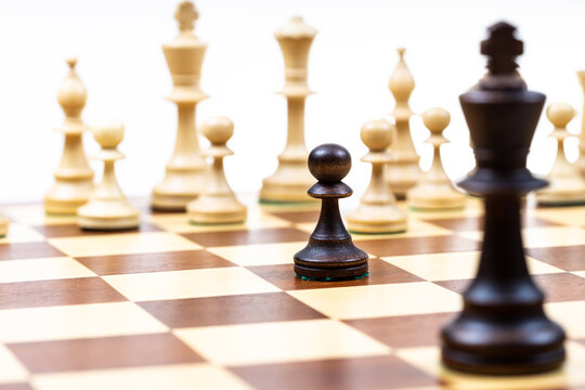 black pawn and single queen against white chess pieces in background on wooden chessboard close up (focus on the black pawn)
