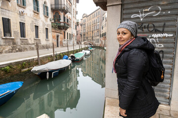 Fototapeta na wymiar A young backpacker woman in winter clothes visiting Venice's canals. Boats on water and graffiti on the wall