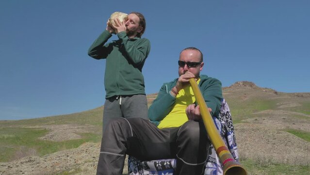 Guys play music on didgeridoo and sea shell. Friends are outdoors