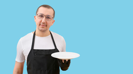 Portrait of a waiter in a black apron holding an empty plate and looking at the camera on a blue background. Cooking concept