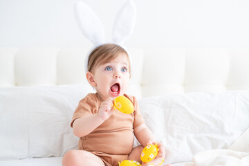 cute funny baby with bunny ears and colorful Easter eggs on bed at home