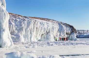 Frozen Baikal Lake. In winter, coastal cliffs of Olkhon Island are covered with white crust of splashing ice with long icicles and bizarre ice grottoes. Group of tourists inspecting scenic icy rocks
