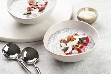 Breakfast idea - two bowls with with chia seeds pudding, red raspberry smoothie, pecan nuts, coconut flakes and pepitas on gray background