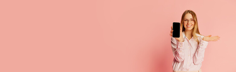Fototapeta premium Happy young pretty blonde girl, student wearing modern office style clothes showing phone screen over pink background. Banner with copy space