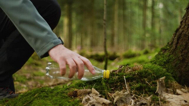 Unrecognizable man picking up a discarded empty plastic water bottle in a forest in Europe. Real time, shallow depth of field, low angle view