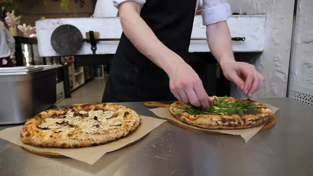 A close-up of a chef decorating a freshly baked pizza with arugula in the pizzeria kitchen against the background of an Italian traditional oven.