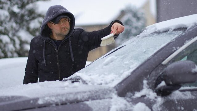 Cleaning snow from windshield of SUV car with brush during heavy snowfall cyclone. Snowstorm in winter season. Vehicle care in blizzard