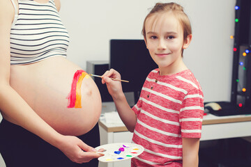 Cute boy drawing rainbow on pregnant belly his mother. Baby birth expecting time and belly painting.