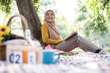 Photo of muslim girl student wearing headscarf sitting on blanket in green park and reading book...