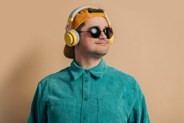 Stylish caucasian man in green shirt, headphones, sunglasses and cap on brown background