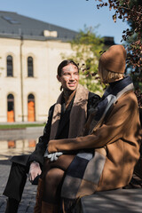 Naklejka premium happy tattooed man looking at blonde woman in hat while sitting on bench.