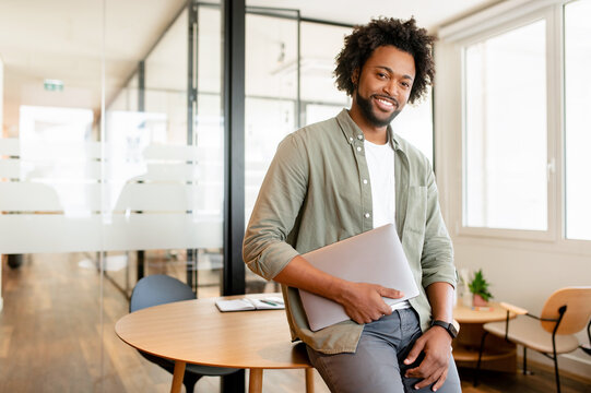 Portrait of confident African-American businessman standing with laptop, male office employee in smart casual wear looking at the camera and smiles, leaned back at te desk, ceo, manager, sturtup owner