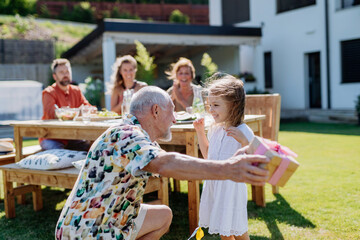 Happy little girl giving birthday present to her senior grandfather at generation family birthday party in summer garden.