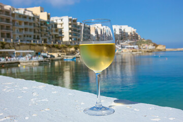 Close up view of glass of white wine with beautiful Malta island of Gozo coast resort town Marsalforn on background with blue sky and sea. Lot of copy space on blur background.