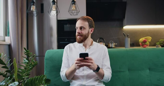 Handsome man browsing smartphone while sitting on sofa in kitchen at home. Cheerful bearded caucasian male in white shirt is texing and read a message.