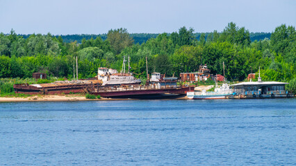 Old rusty ships lie on the riverbank. The river bank and rusting ships. Pier with a boat and old...