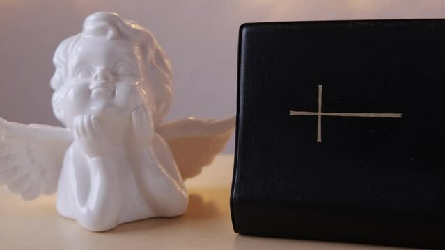 female hand leafs through pages of old black book, family bible in dark cover, white ceramic figurine of angel with wings, concept of intermediary between God and world, all saints' day