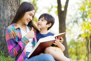 Fototapeta na wymiar Happy cheerful Asian lovely little boy and mother sitting under the tree at park in the morning and reading a book together. Asian woman spending time in weekend with her lovely little son.