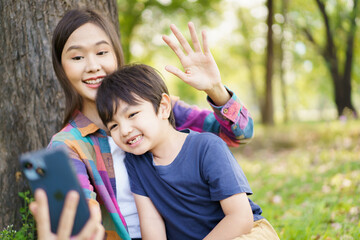Happy cheerful Asian little boy using smartphone to take a selfie photography with his mother at the park in morning. Happy family time concept, woman staying with her little son in weekend.