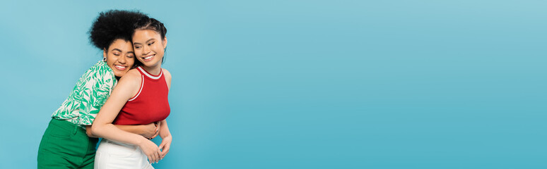 joyful african american woman with closed eyes hugging smiling asian friend isolated on blue, banner.