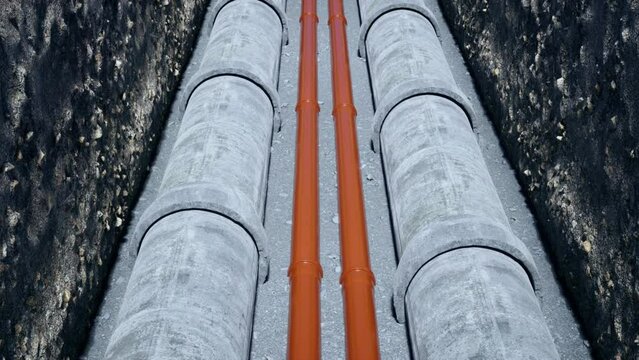 An aerial view over a newly dug trench containing precast concrete pipes and bright orange plastic pipes, installed for the construction of a new housing development.