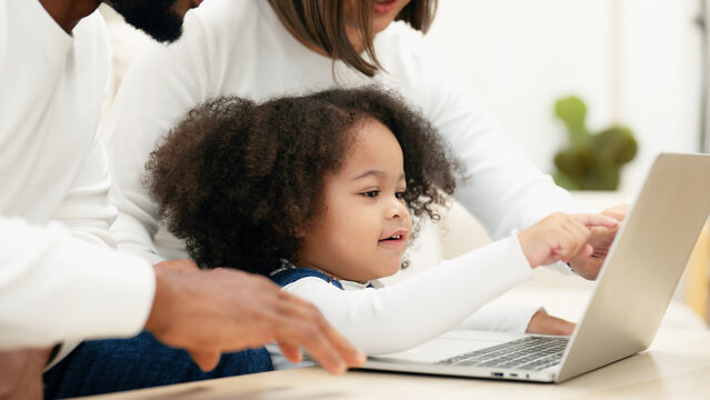 Close-up of a curious daughter touching a laptop screen to select online kid contents while multiracial newbie parent taking care of her. A happy family is teaching their daughter to use a laptop.
