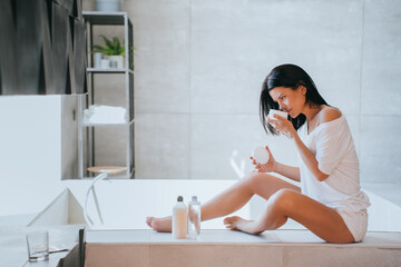 Beautiful brunette hispanic young adult woman sitting at bathroom smells new skincare cream, spa procedure. Caucasian housewife trying different lotions for legs, moisturiser. Health care.