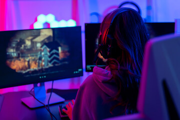 Back of a woman with confidence is going live or streaming during a game. She wears headphones and speaks to her teammates or those who are watching her on social networks or online platforms.