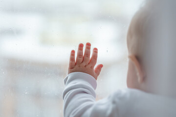 Naklejka premium BabyToddler is standing with his hand on the window and looking at the street.