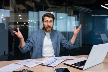 Portrait of disgruntled financier businessman inside office, male angry boss looking at camera and shouting displeased with report and paid bills, man doing paperwork inside office with laptop. - obrazy, fototapety, plakaty