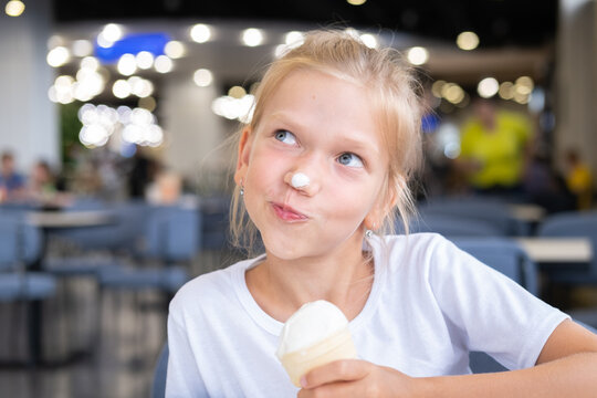 Portrait of a little hungry funny girl eating cold delicious ice cream in a waffle cup sitting in a cafe, dark background, bokeh. A child enjoys ice cream. A sweet snack