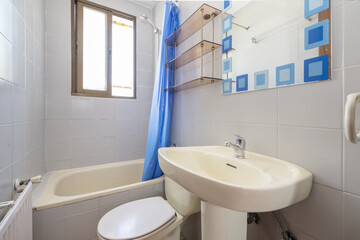 Small bathroom with aluminum windows, a frameless mirror with blue patterns, a porcelain sink with a porcelain pedestal, and a small bathtub with a blue curtain