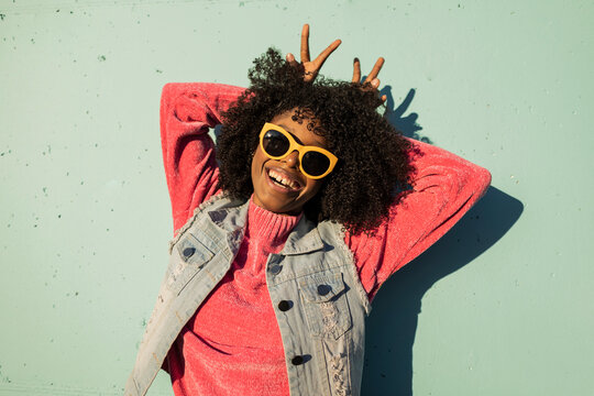 Cheerful young woman wearing sunglasses showing peace gesture in front of wall