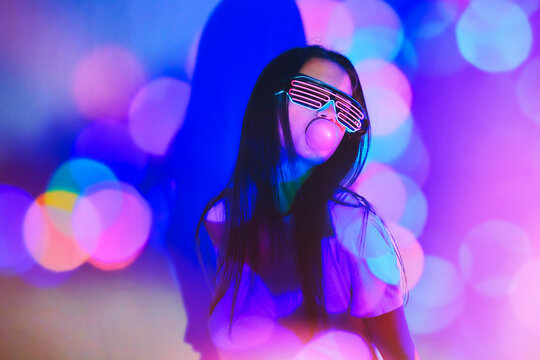 Young woman with futuristic glasses blowing bubble against neon colored background