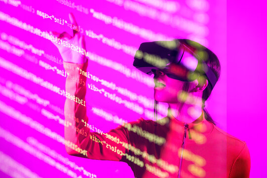 Woman with VR goggles touching computer codes over pink background